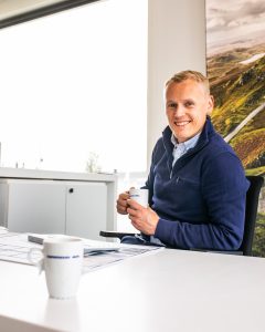 Mann i blå genser sitter bak en hvit kontorpult med en kaffekopp i hendene